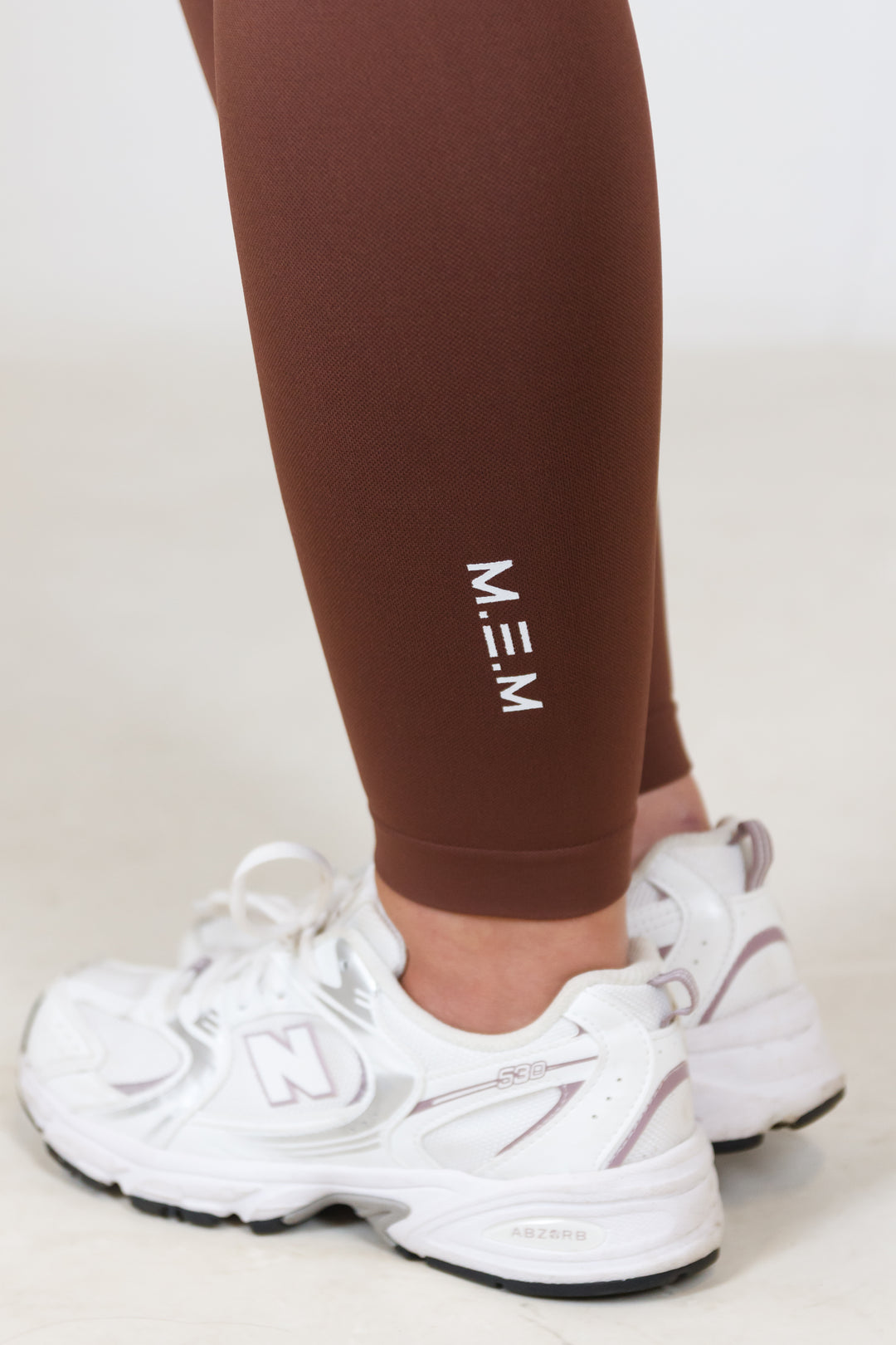 Brown leggings with 'MEM' branding worn by a person, paired with white sneakers on a light background.