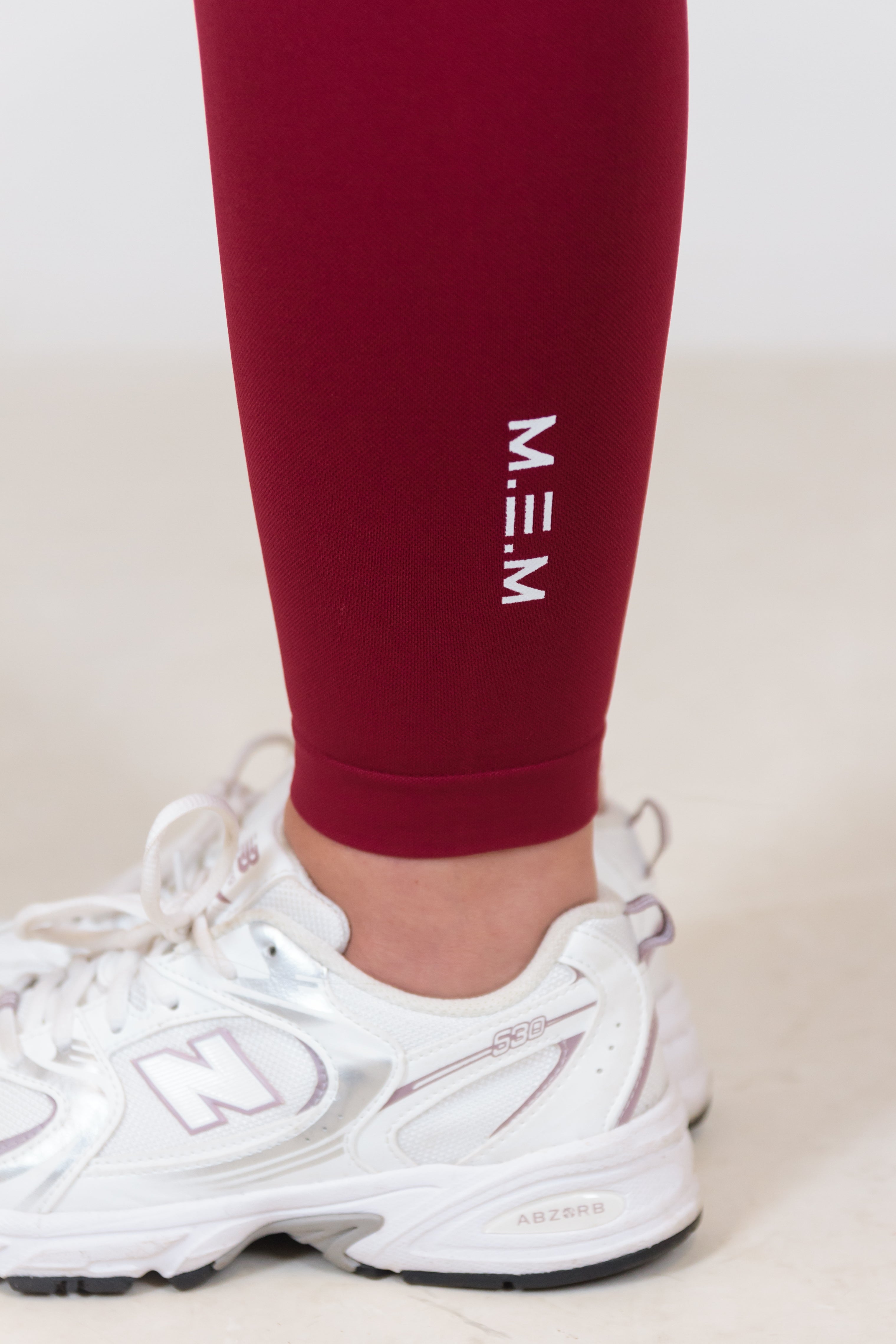 Maroon leggings with 'M.E.M' logo worn by a person, paired with white sneakers on a light background.